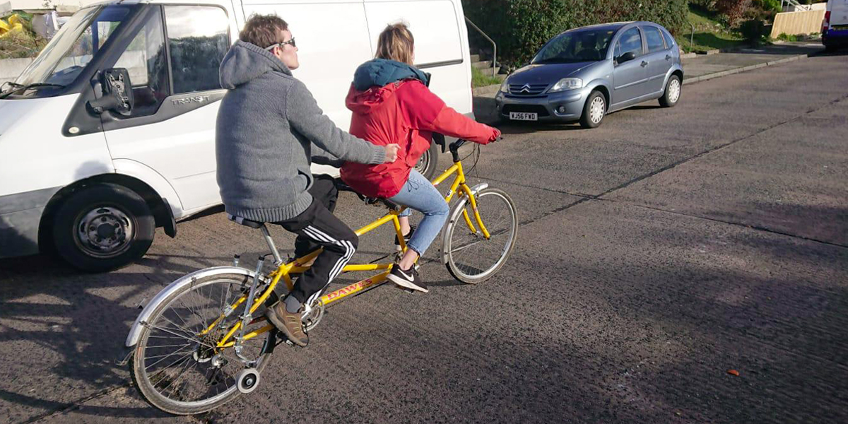 Photo of Ricky and his support worker riding a tandem.